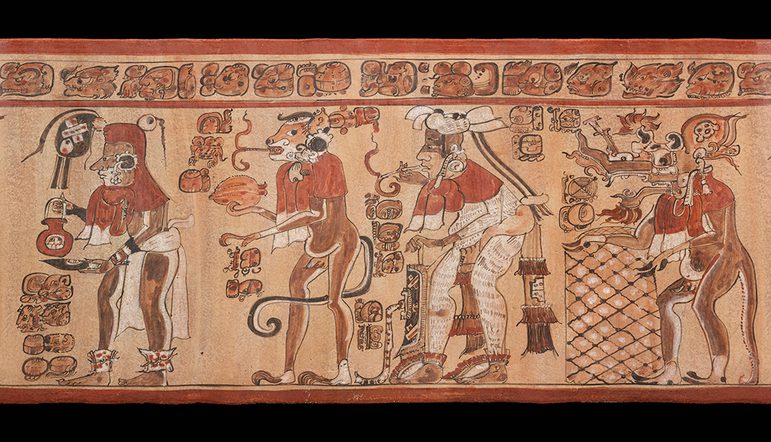 Ancient Mayan vase depicting elaborately dressed figures engaged in a ritual. Each figure is adorned with intricate headgear and jewelry, and holds ceremonial objects. A detailed hieroglyphic script runs above the scene.