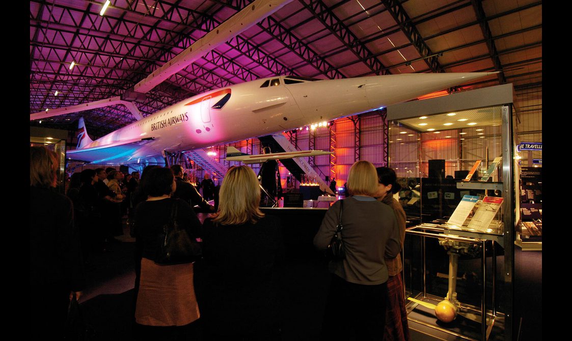 People stand grouped around tables under the Concorde, which is lit with purple and blue lights.