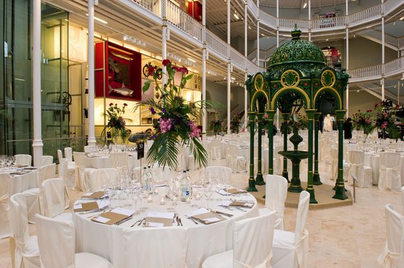 A large, bright, multistory space with balconies running around the sides. The floor is filled with large round tables covered in white tablecloths and set for a meal. The tables are surrounded by white chairs.