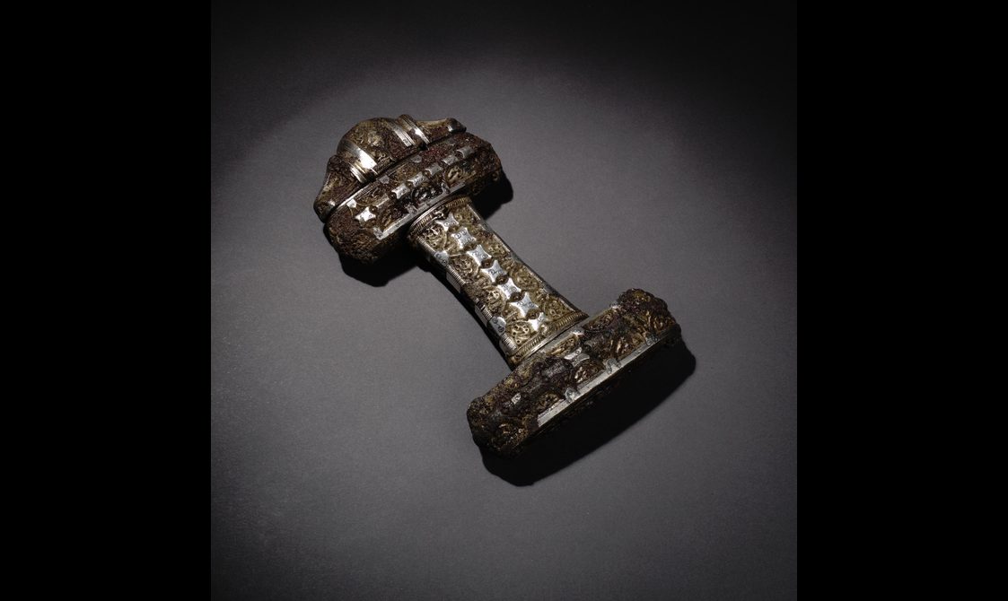 A sword hilt assembled from three rectangle parts in a shape of a horizontal letter H with a rounded triangle-shaped part at one end. Inlaid with non-descriptive silver organic shapes throughout.