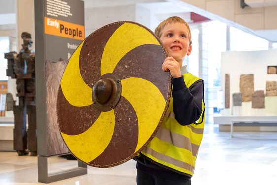 A child in a high-vis vest stands in a gallery space holding a large, round, gold and yellow shield.