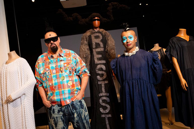 Two people stand in front of mannequins wearing black and white clothes. One of the people wears a colourful shirt and sunglasses, the other wears a navy dress and has bright blue makeup around their eyes