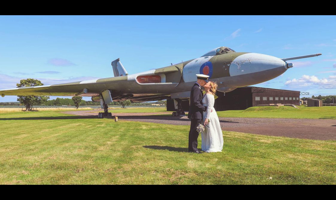 A man in a black suit and white military hat kisses a woman in a white wedding dress. They are standing on the grass in front of the Vulcan airplane on a bright day with a blue sky.