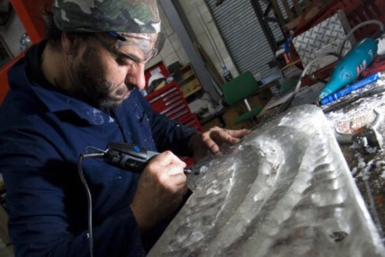 A man wearing navy overalls and a bandanna carving into a decorative piece of wood in a workshop