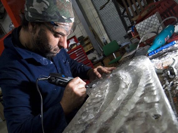 A man wearing navy overalls and a bandanna carving into a decorative piece of wood in a workshop