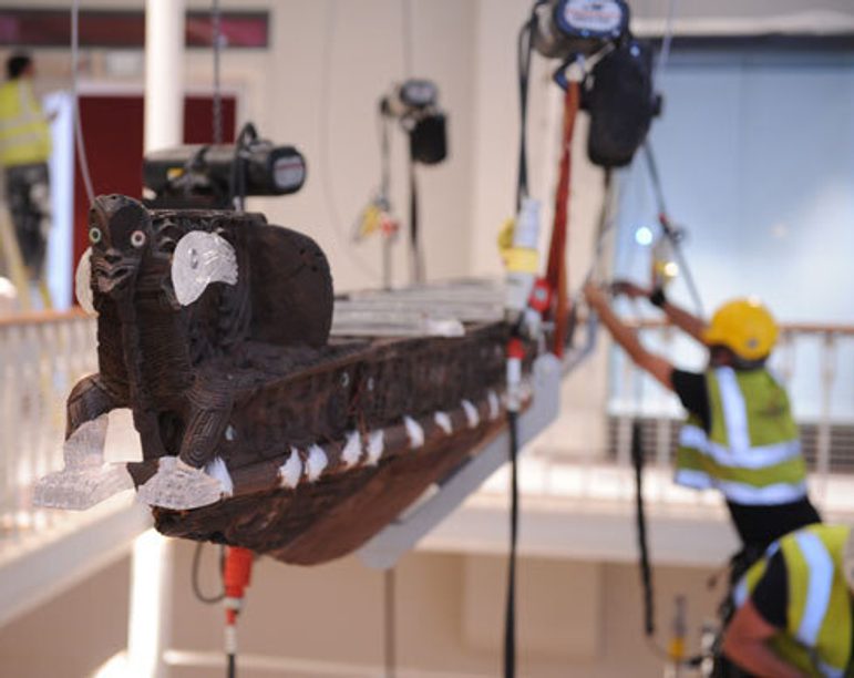People in high-vis vests and hardhats suspending a wooden and perspex canoe from the ceiling with pulleys and ropes