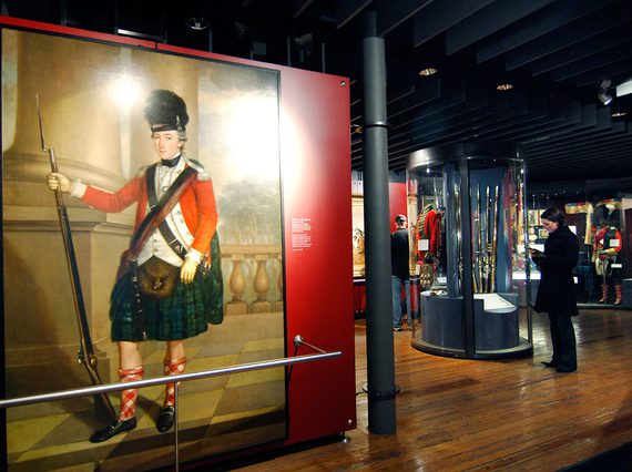 A museum gallery with a large portrait of a Scottish soldier in traditional uniform dress. There are visitors in the background looking at objects in cases.