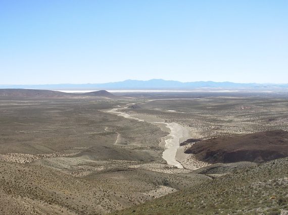 A gently rolling landscape from above covered in scrub bushes with large mountains in the distance.
