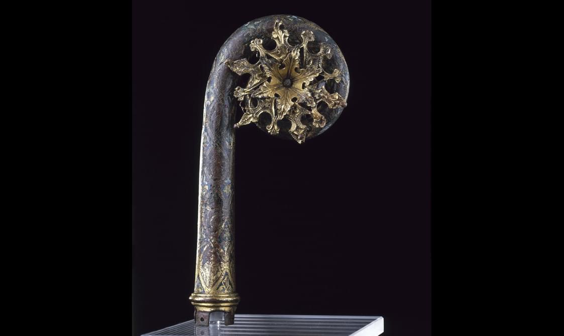 Metalwork object with a staff and a coiled top. The staff is grey-blue, and a golden sun-like burst is within its coiled top.