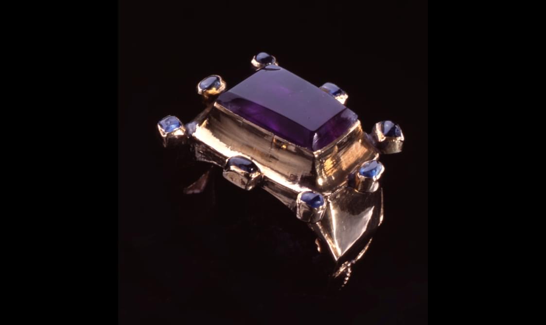 Ornate thick gold ring with a large rectangular purple gemstone surrounded by eight smaller purple gemstones.