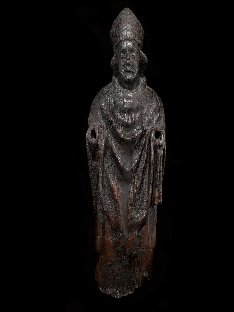 Wooden figure of a saint in flowing robes. The figure is heavily worn and darkened, its arms missing and its eroded face seems melancholic.