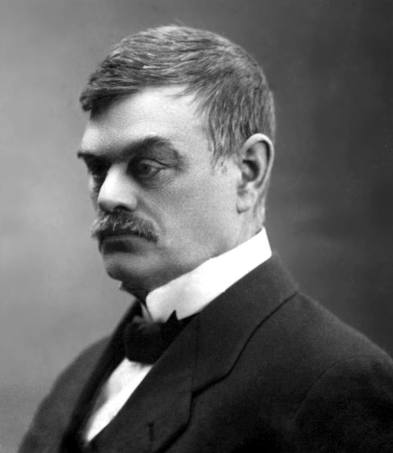 A black and white portrait of a man from the shoulders up. He has short hair and a mustache, and is wearing a white shirt with a bow tie and jacket.