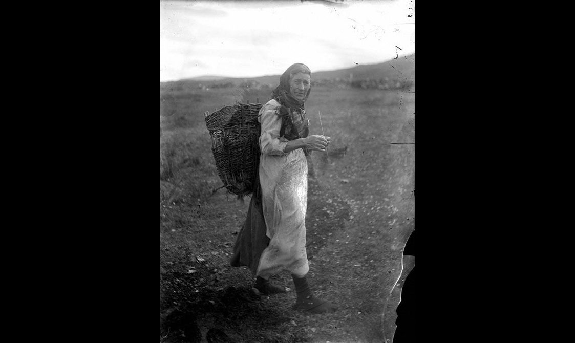 A black and white photograph of a woman wearing a long white dress with a shawl around her head, carring a basket on her back while knitting as she walks through the countryside.