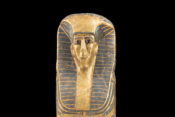 A gold and black coffin of an Egyptian woman photographed against a black background. The coffin features a female face wearing a headdress.