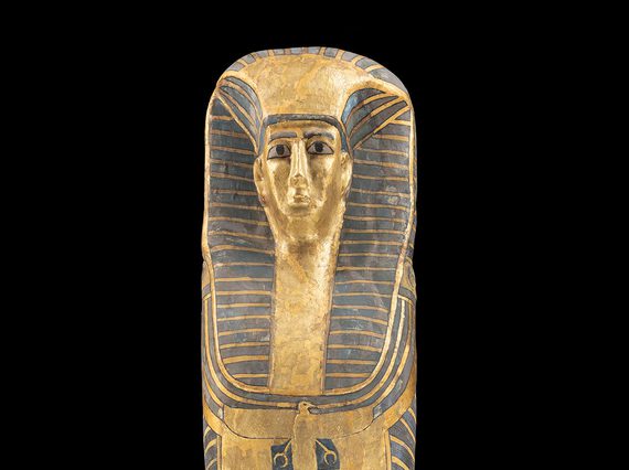 A gold and black coffin of an Egyptian woman photographed against a black background. The coffin features a female face wearing a headdress.