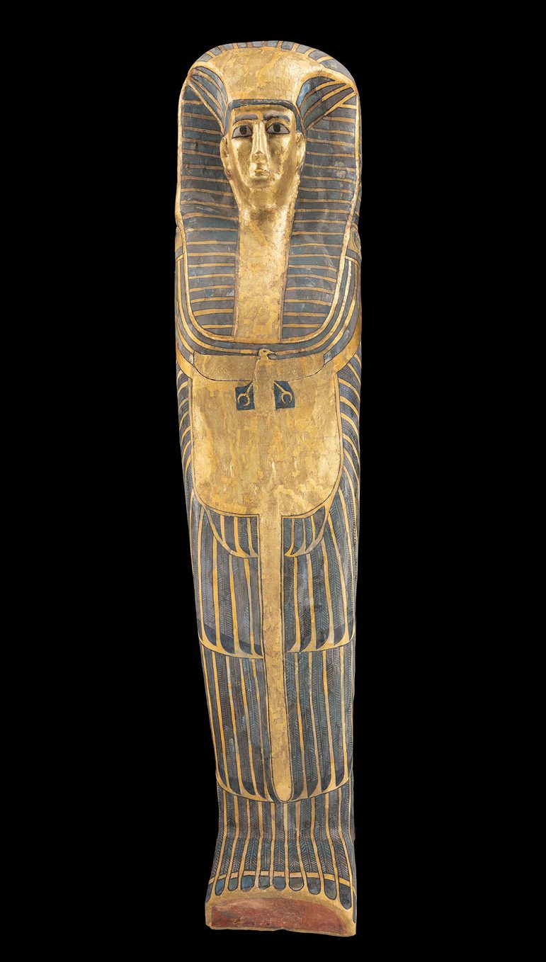 A gold and black coffin of an Egyptian woman photographed against a black background. The coffin features a female face wearing a headdress.