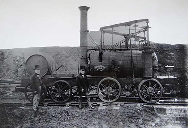 A black and white photograph of two men in Victorian suits and top hats beside a steam engine train.