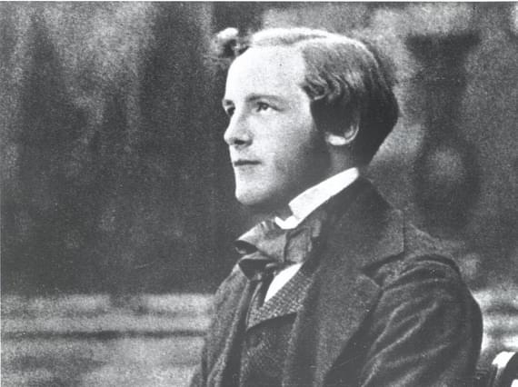 A black and white photograph of a young man wearing a white collared shirt, dark suit jacket and waistcoat, sitting on a bench holding a spinning top.