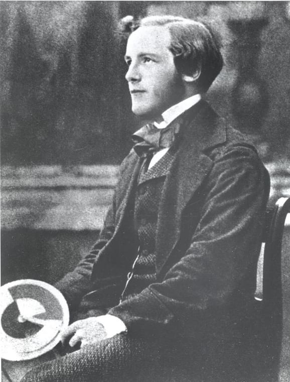 A black and white photograph of a young man wearing a white collared shirt, dark suit jacket and waistcoat, sitting on a bench holding a spinning top.