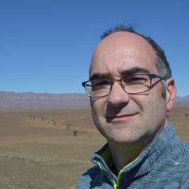 A head and shoulder portrait photo of a man with salt-and-pepper hair, wearing blue framed glasses and a marled blue fleece. He is standing in the field at the world-famous Fezouata fossil locality in Morocco, with in the background a blue sky and the hills of Jbel Rhart.