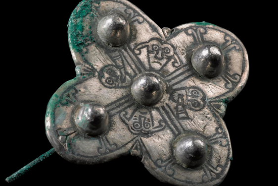 A tarnished silver brooch in a clover shape with raised balls on each point and in the centre. There are simple line drawn faces etched into its surface